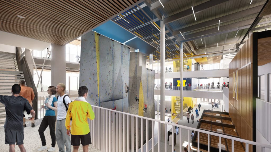 Indoor facility with a tall rock climbing wall, groups of people walking and talking, and several upper floors visible with additional activity.