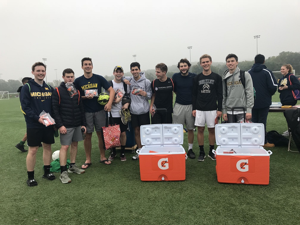 Group of students on a grassy athletic field, posing with drinks and snacks behind two open orange Gatorade coolers.