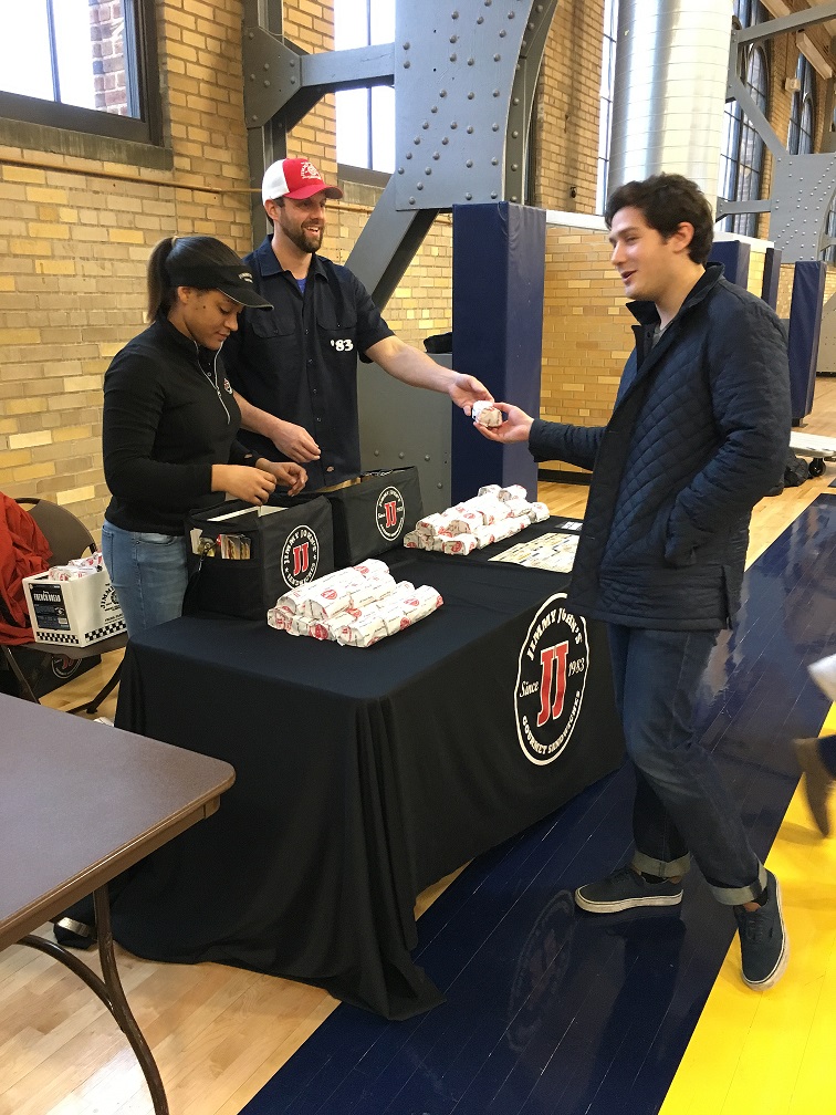 Table with wrapped sandwiches and staff handing food to a person.