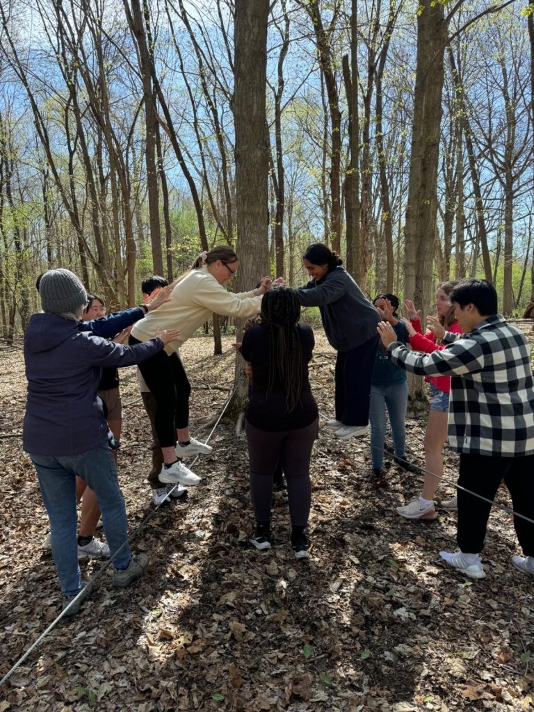 People navigate a low wood balance beam in a forest, working together for stability while another person observes