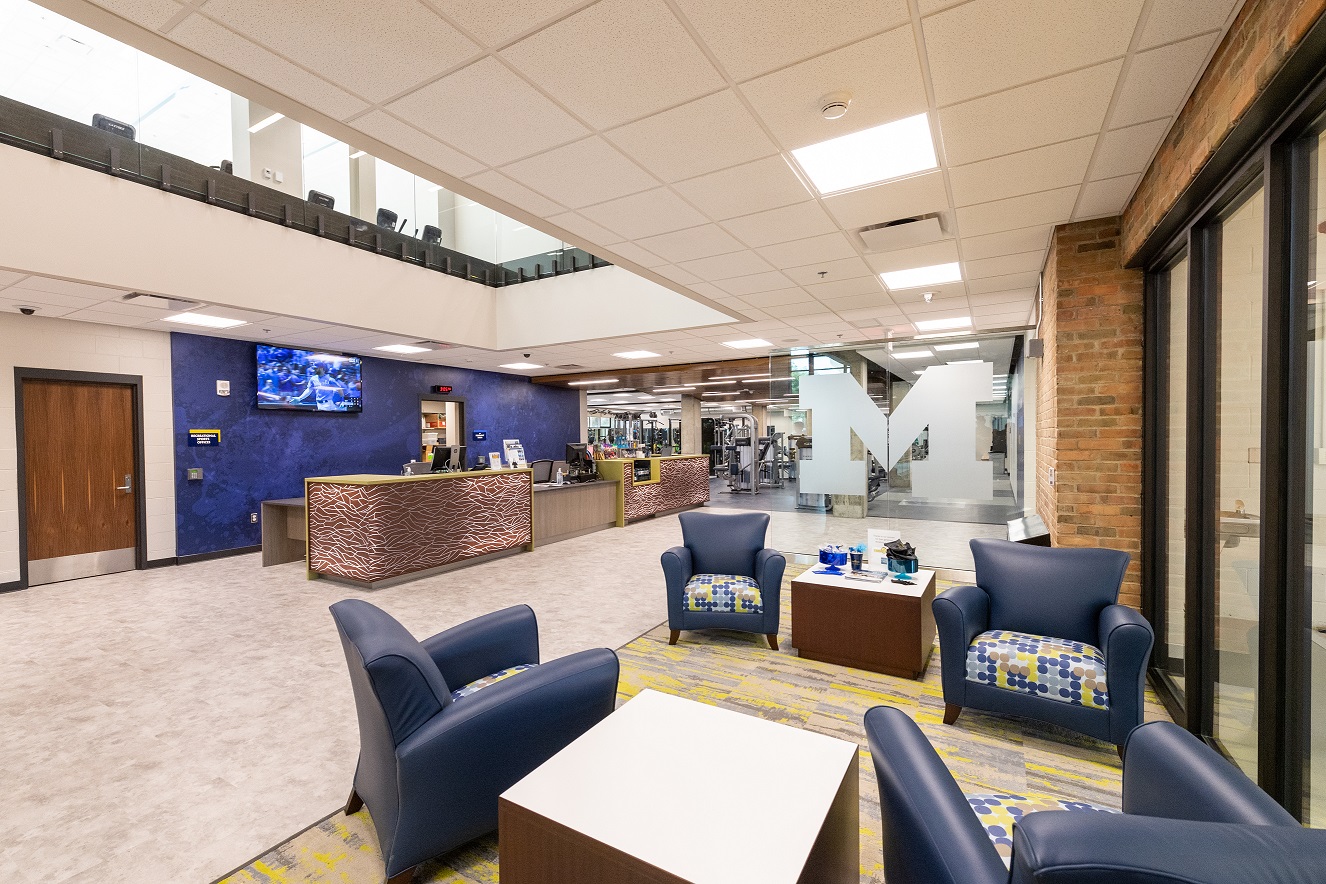 Comfortable seating area with blue chairs, lobby desk, and Michigan logo.