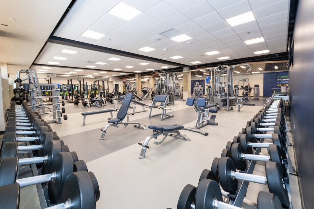 Gym with rows of dumbbells, adjustable benches, strength machines, and mirrors throughout.