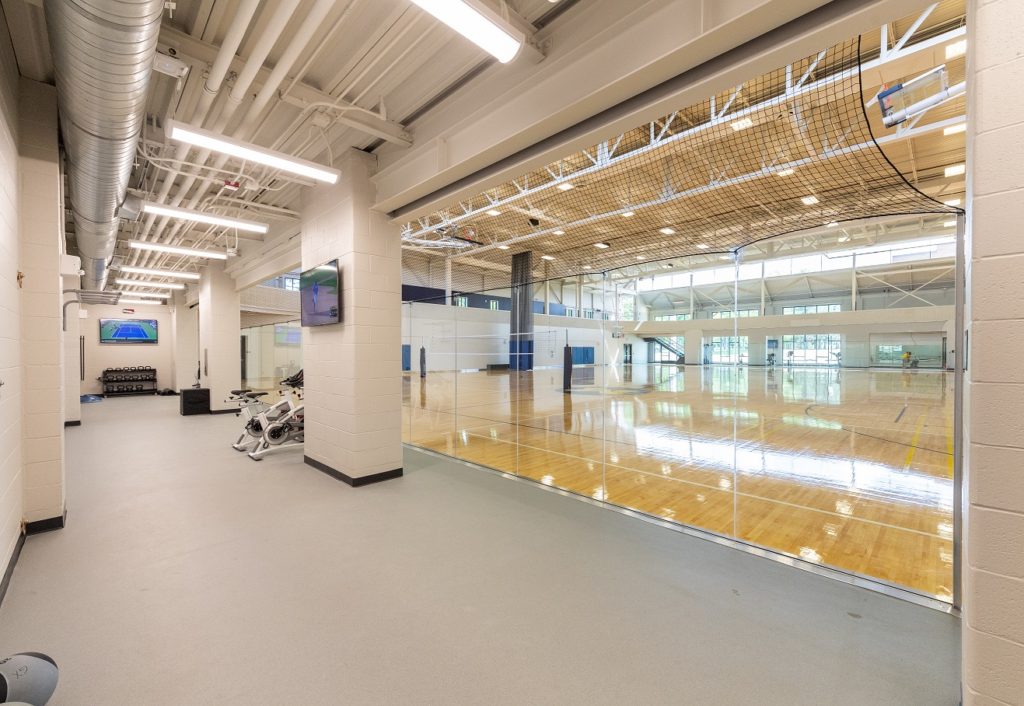 Fitness area with stationary bikes alongside a glass wall, view into indoor basketball court with wooden flooring and high ceilings.