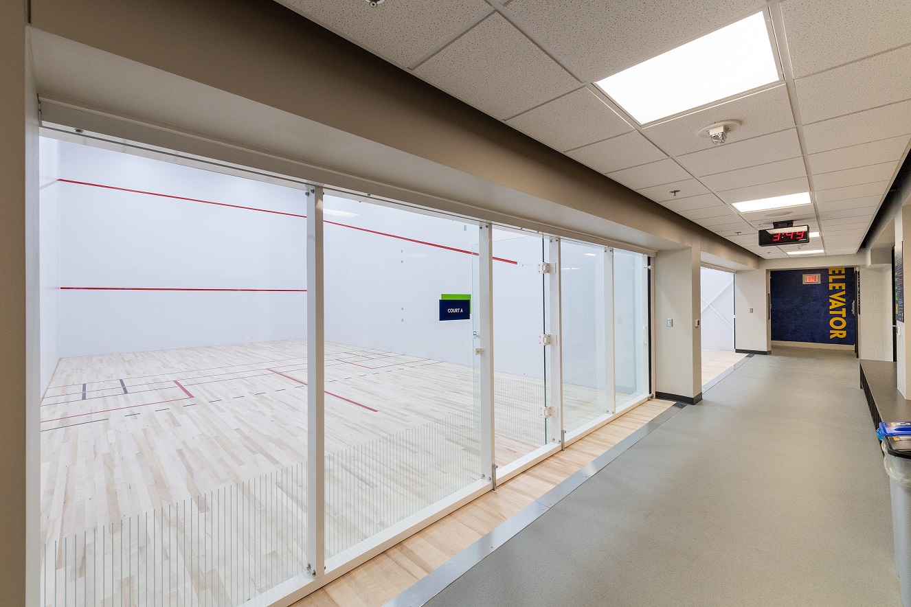 Indoor squash court with glass walls, wooden floors, red court lines, and hallway lighting.