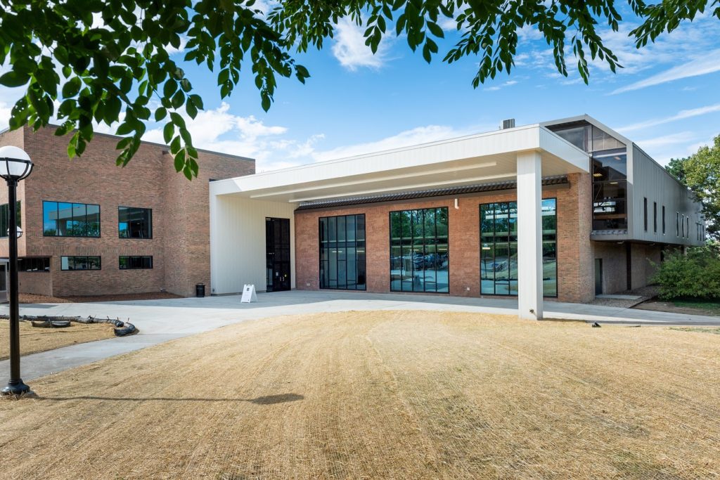 Modern campus structure with brick walls, white awning, glass windows, and open, grassy entrance area beneath tree branches