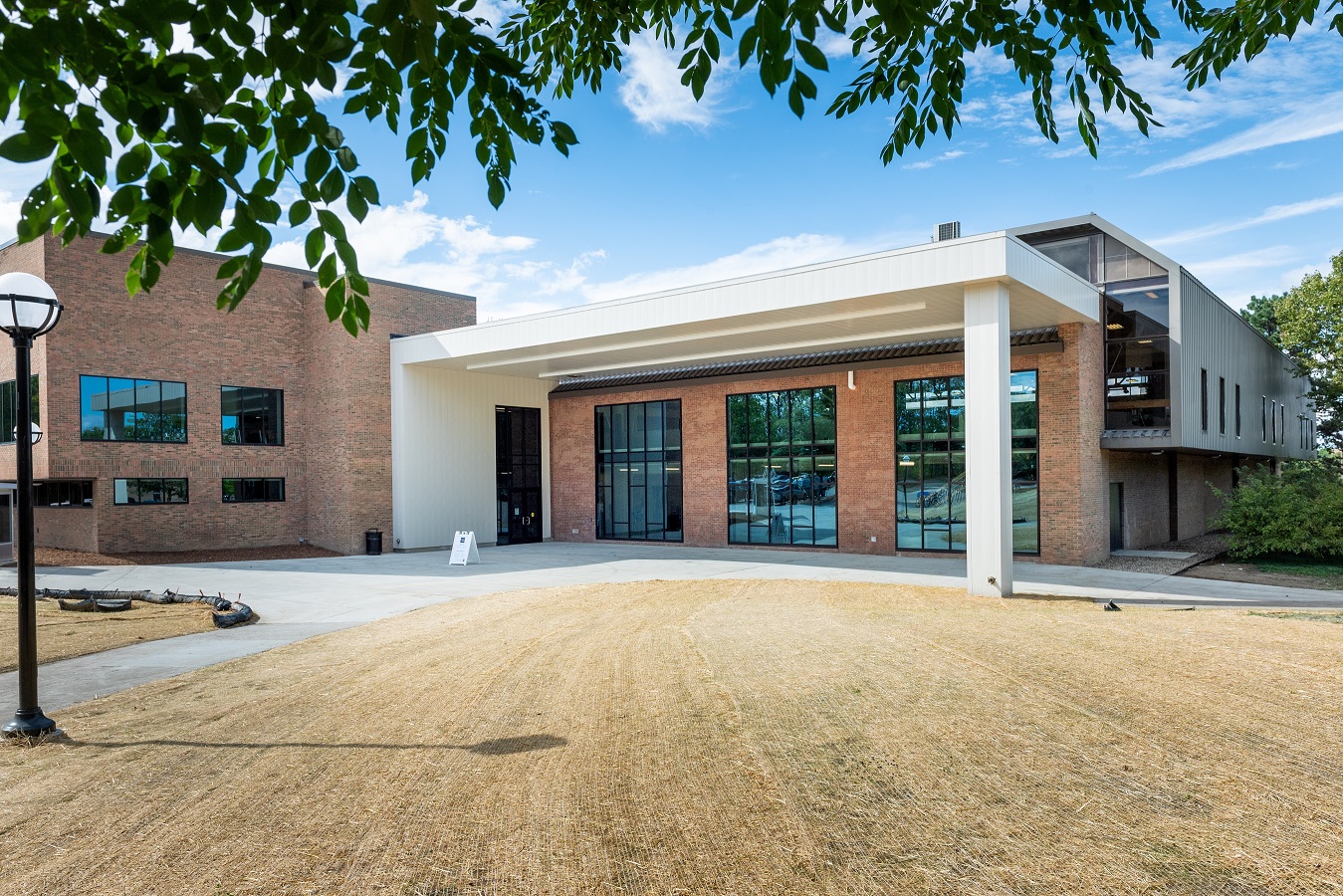 Modern campus structure with brick walls, white awning, glass windows, and open, grassy entrance area beneath tree branches