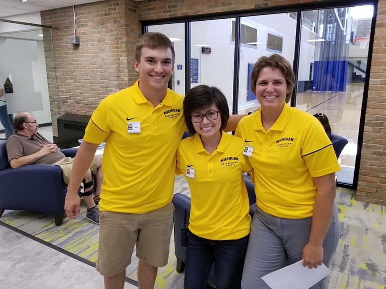 Three people in yellow Michigan staff shirts pose together indoors.