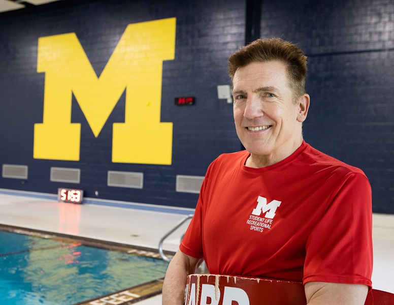 Lifeguard in red shirt stands poolside near large maize block “M.”