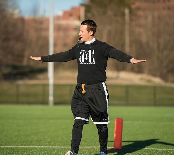Referee in black Michigan shirt stands with arms outstretched on grassy field.