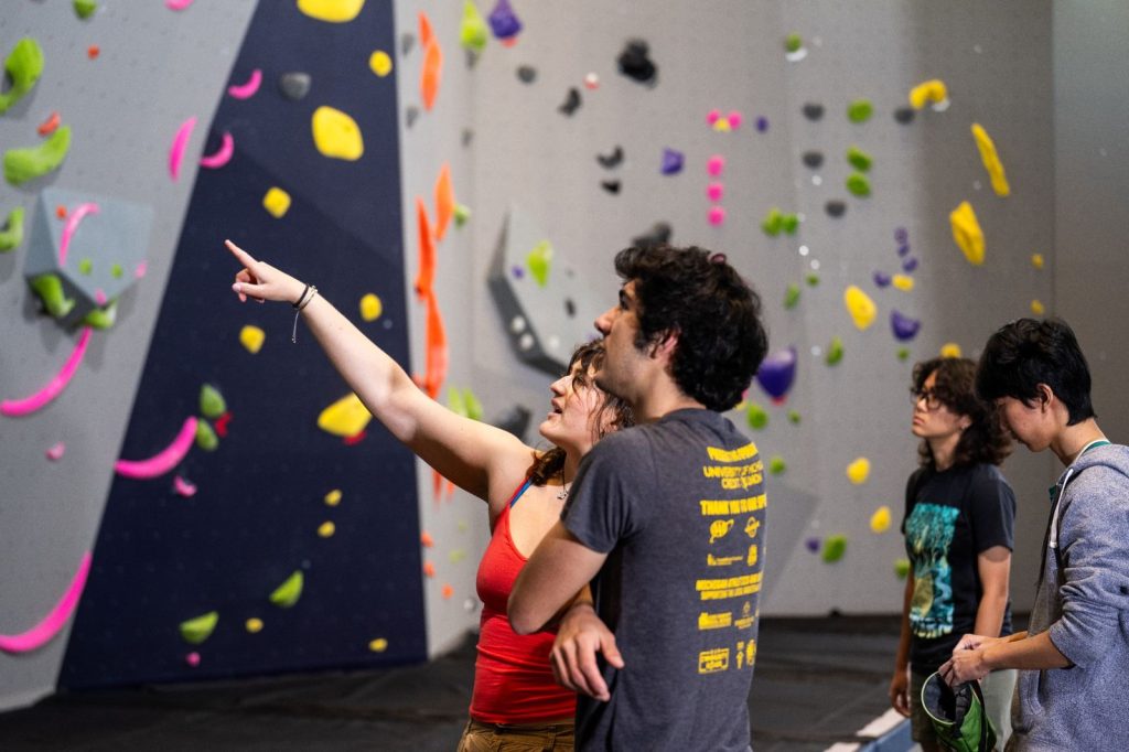 Two people looking at an indoor climbing wall, with one person pointing toward the climbing holds