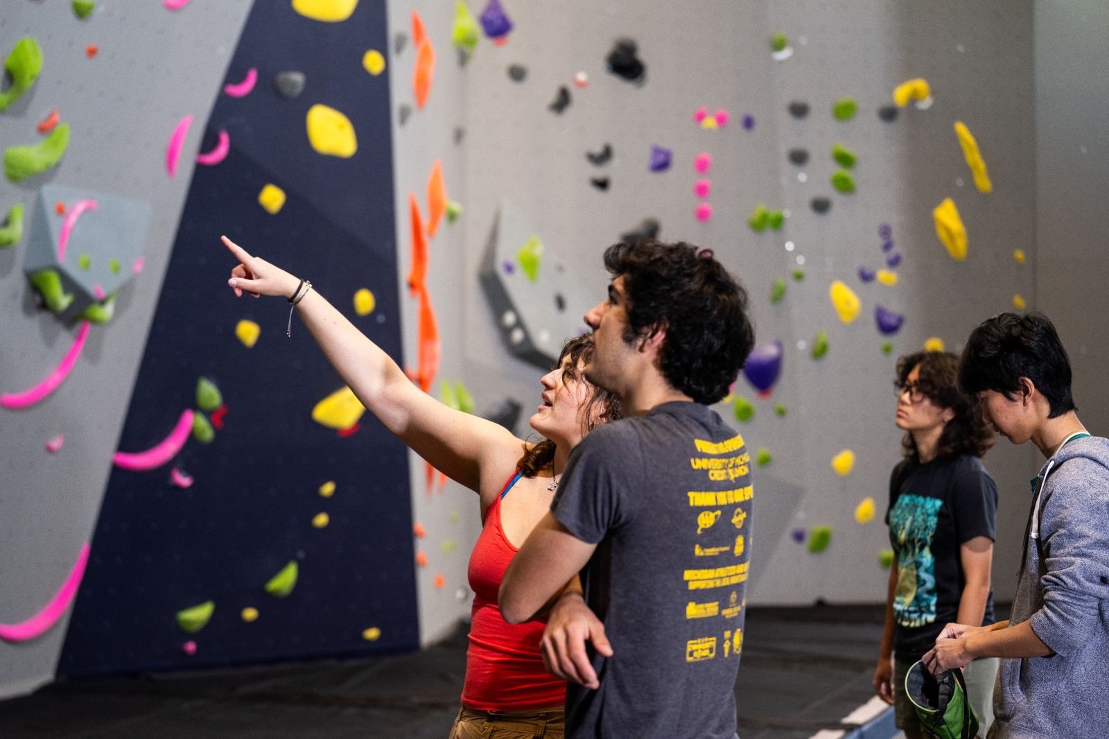 Two people looking at an indoor climbing wall, with one person pointing toward the climbing holds