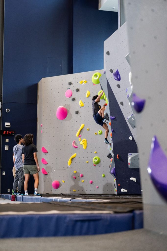A person climbing a colorful indoor rock wall while two people stand nearby