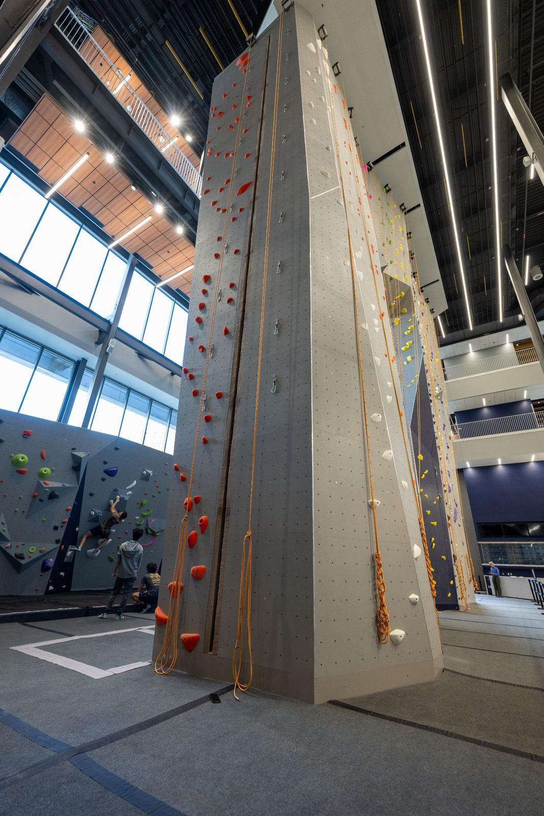 Tall indoor climbing walls with red climbing holds and hanging ropes, with climbers in the background