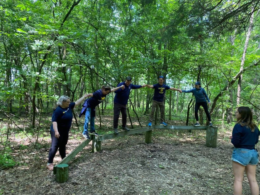 Group balances on wooden beam in forest, supported by peers.